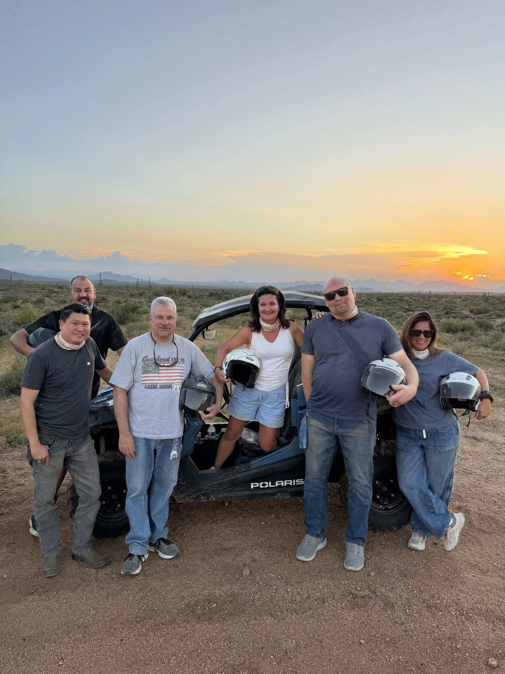 A corporate team of six holding helmets beside a Polaris RZR at sunset on a Scottsdale desert team outing