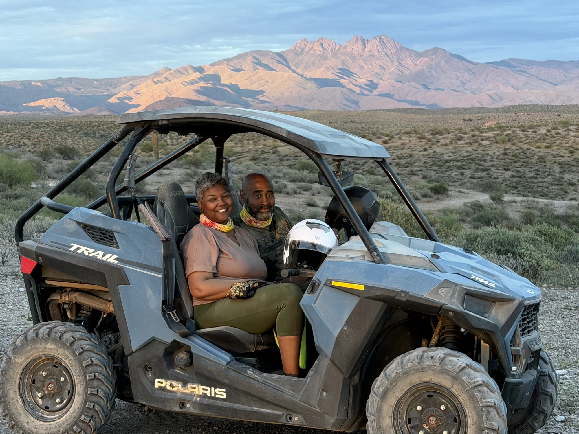 A smiling couple seated in a Polaris RZR Trail as pink alpenglow lights up the Four Peaks on a Scottsdale sunset UTV tour