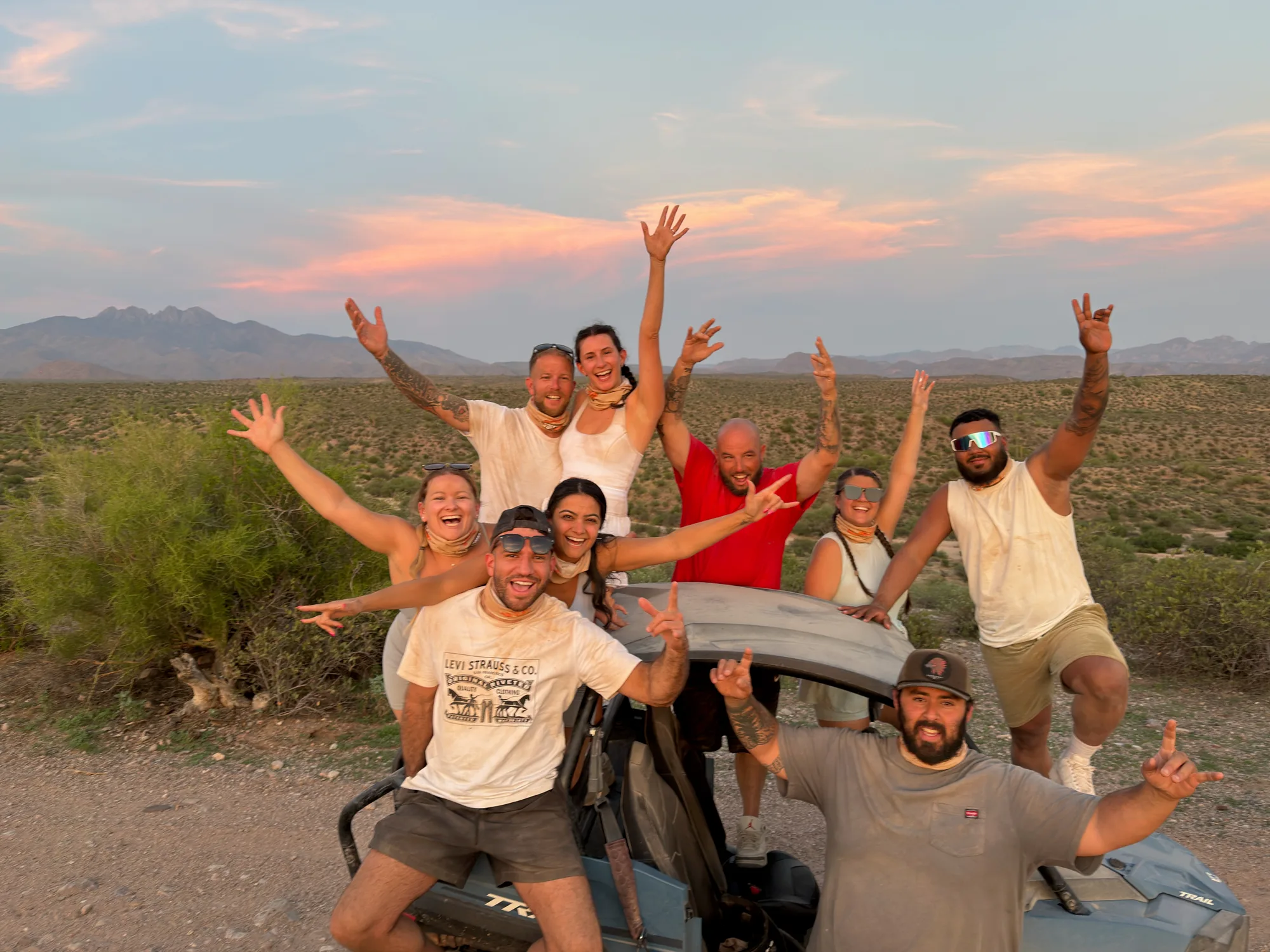 A cheering tour group with arms raised around a Polaris RZR at sunset below the Four Peaks near Scottsdale
