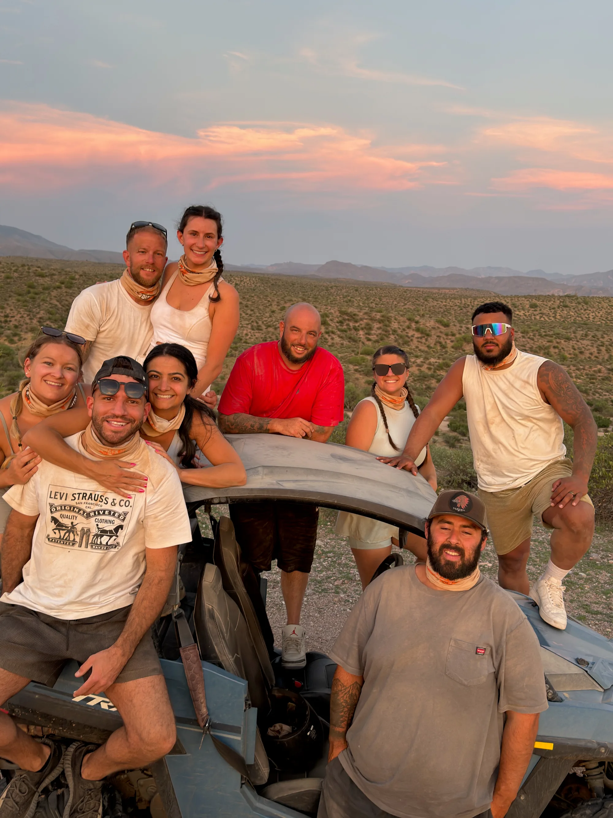 A group of friends gathered around a Polaris RZR under a pink sunset sky on a Scottsdale desert tour
