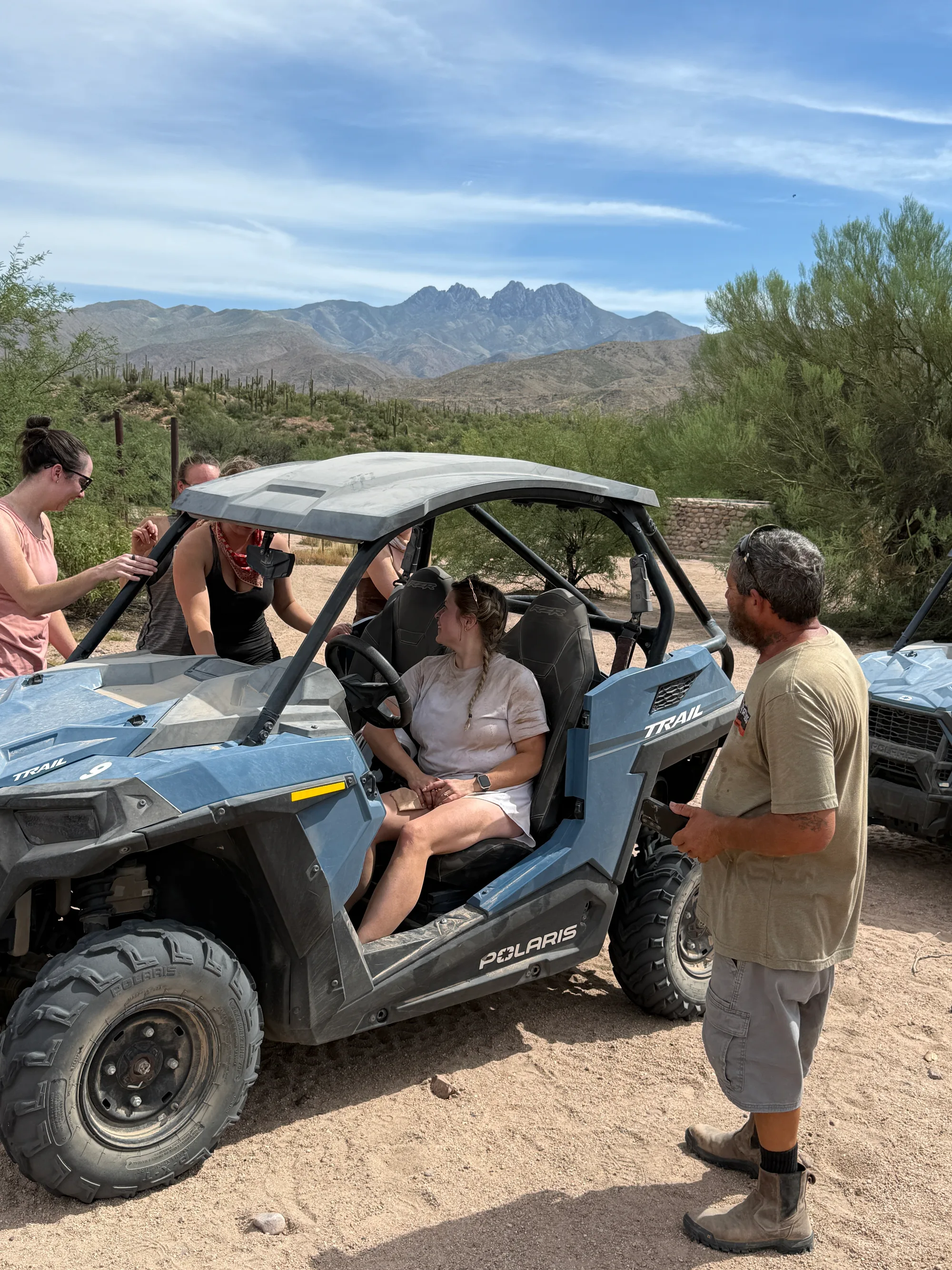 A Desert Dog Offroad Adventures guide giving a rider her safety briefing in a Polaris RZR with the Four Peaks behind