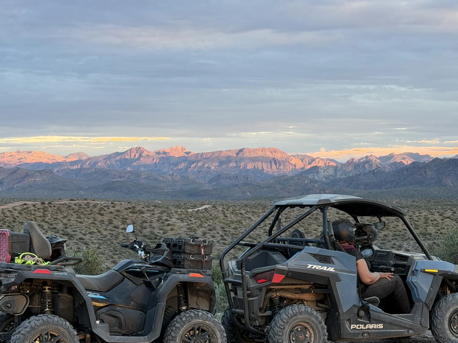 An ATV and Polaris RZR parked on a desert ridge as alpenglow lights the mountains at dusk near Scottsdale